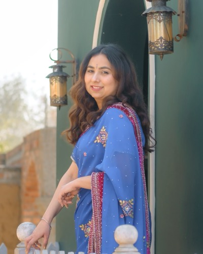 Navneet Kaur, smiling, in a blue saree at an arched green doorway between two lanterns